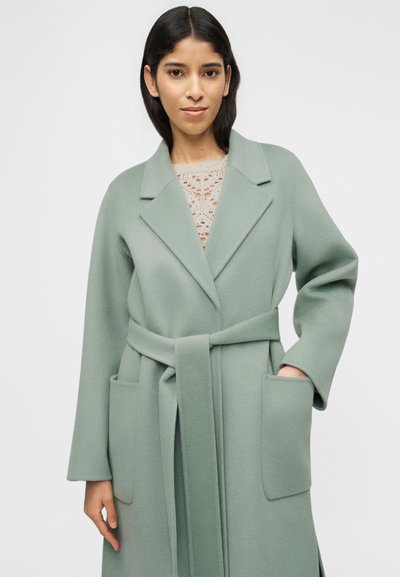 Γυναίκα που φοράει παλτό από μάλλινη ύφανση σε απόχρωση φασκόμηλου με ζώνη, από πάνω από δαντελωτό τοπ, στέκεται με το ένα χέρι στην τσέπη του παλτού μπροστά σε απλό ανοιχτό φόντο.