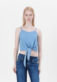 Top corto in denim blu chiaro con sottili spalline a spaghetti, scollatura a V e dettaglio con nodo anteriore. Indossato con jeans blu scuro.