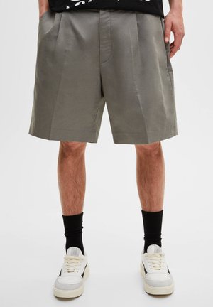 Szare szorty wykonane z gładkiego materiału, z plisowanym wzorem i luźnym krojem, połączone z białymi trampkami i czarnymi, prążkowanymi skarpetkami.