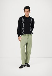Zwarte lange mouwen polo met witte bloemenborduursels, gecombineerd met lichtgroene baggy broek en zwarte schoenen met witte strepen. Eenvoudige achtergrond.