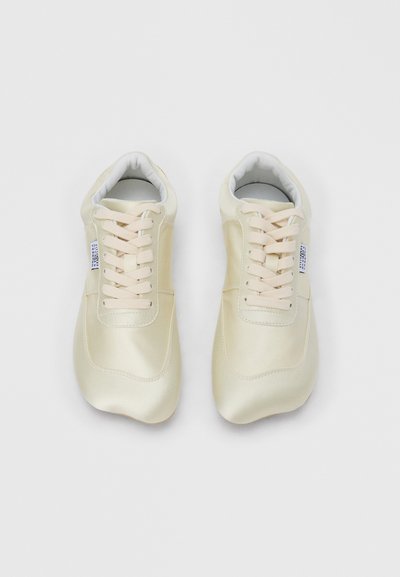 Ζευγάρι κρεμ παπούτσια αθλητικά με κορδόνια και μικρή λευκή ετικέτα στο πλάι, απεικονισμένα από πάνω σε λευκό φόντο.