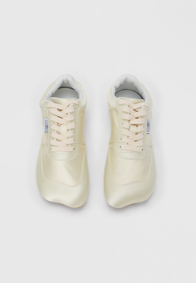 Par cremefarvede snøresneakers med en lille hvid mærkat på siden, vist fra en top-down vinkel på en hvid baggrund.