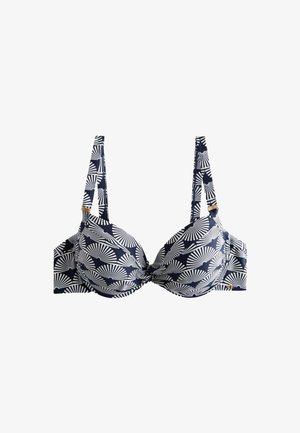 Frau mit langen dunklen Haaren, die einen marineblau-weißen Bikini mit Fächer-Muster trägt, steht vor einer beigen Wand im natürlichen Licht.