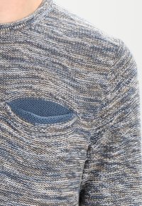 Pull en maille avec un motif texturé dans des tons de bleu et de beige. Comprend un détail de poche bleu distinct sur le devant à gauche.