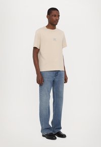 Beige bomulds T-shirt med korte ærmer, med et lille blåt logo på brystet. Sæt sammen med lyseblå lige ben jeans og sorte sko.