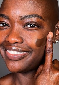 Foundation påført huden, rik brunfarge, glatt tekstur. Modells hånd med lys neglelakk peker på foundation på kinnet.