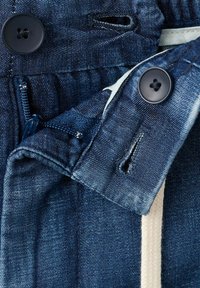 Gros plan d’un vêtement en jean bleu foncé déboutonné montrant des boutonnières, des boutons noirs et une fermeture éclair partiellement visible avec un tissu blanc à l’intérieur.