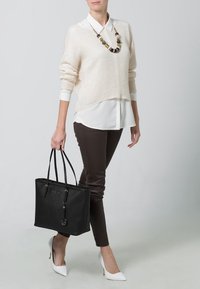 Sac fourre-tout noir avec deux poignées, finition texturée et un charme circulaire. Porté avec un pull en tricot beige, une chemise blanche à col et un pantalon marron.
