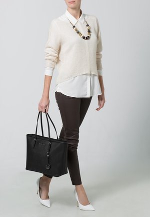 Sac fourre-tout noir avec deux poignées, finition texturée et un charme circulaire. Porté avec un pull en tricot beige, une chemise blanche à col et un pantalon marron.