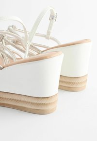 Sandales compensées avec tige blanche, plateforme beige texturée, brides argentées scintillantes et boucle réglable à la cheville. Design de talon arrondi.