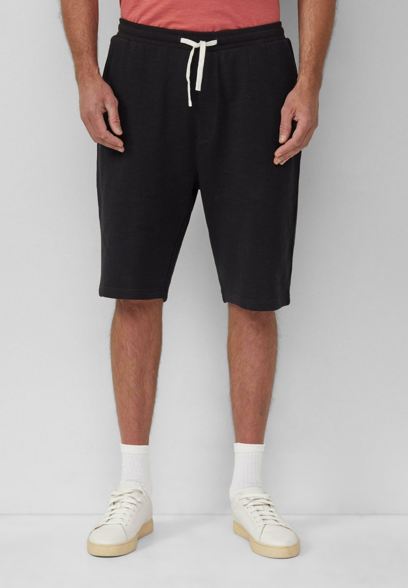 Schwarze Baumwollshorts mit einem Kordelzugbund, Seitentaschen und einem lässigen Schnitt, kombiniert mit weißen Sneakers und Socken.