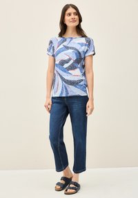 Tópic de manga corta multicolor con patrones abstractos en azul, blanco y rojo, combinado con jeans de pierna recta en azul oscuro y sandalias oscuras.
