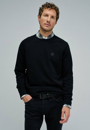 Sweat-shirt noir avec un col rond, des poignets côtelés et une petite étiquette noire. Porté par-dessus une chemise à carreaux, associé à un jean foncé.