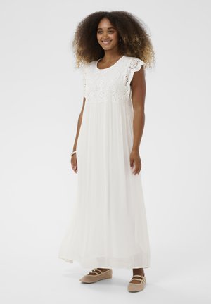 Mosolygó nő, fehér ujjatlan maxi ruhát visel, amelyen kivágott csipke felsőrész található, és bézs lapos cipőt visel, egyszerű fehér háttér előtt.