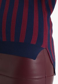 Gros plan sur un pull en maille rayé bleu marine et bordeaux associé à un pantalon en cuir bordeaux, vu de la taille vers le bas.