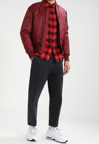 Man in een rode bomberjack, een rood-zwart geblokt overhemd, zwarte broek en witte sportschoenen, staand met zijn handen in zijn zakken.