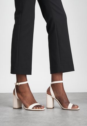 Beige high-heeled sandals with an ankle strap, open toe, and block heel. Smooth material with a matte finish and minimalistic design.