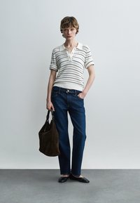 Mladá osoba oblečená v bielej pletenej polokošeli s krátkym rukávom, modrých džínsoch, čiernych balerínach, držiaca veľkú hnedú kabelku, stojaca pred jednoduchou svetlou stenou.