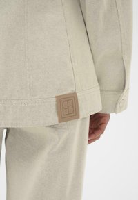 Lys beige jakke med en tekstureret finish, der har et rektangulært logo-patch på bagsømmene og en knaplukning på ærmet.