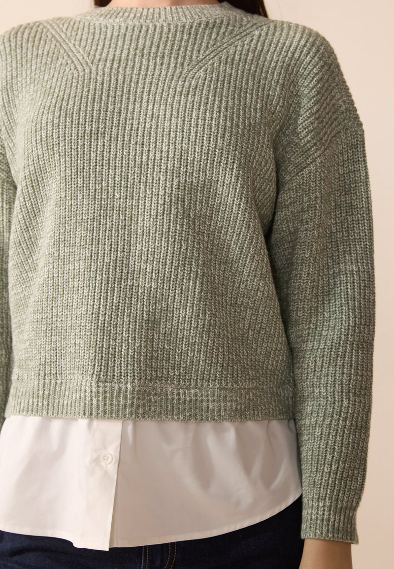 Pull en tricot vert clair avec texture côtelée, col rond et manches longues, superposé à une chemise blanche à col.