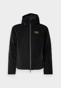 Giacca nera con cappuccio in tessuto trapuntato, dotata di chiusura frontale con zip e di una etichetta con logo dorato sul petto. Design robusto e distintivo.