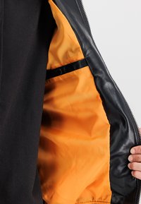 Μαύρο μπουφάν με πορτοκαλί εσωτερική επένδυση; διαθέτει λεία εξωτερική δερμάτινη επιφάνεια, αντίθετη ύφανση στην τσέπη και κλείσιμο με φερμουάρ.