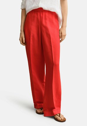 Femme portant un pantalon rouge ample, une chemise blanche à manches longues et des sandales marron, debout devant un fond uni.
