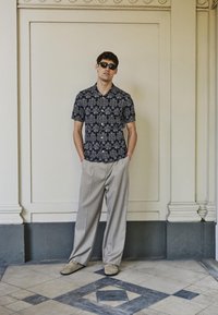 Άνδρας που φοράει μαύρο πουκάμισο με μοτίβο και κοντά μανίκια, ανοιχτό μπεζ παντελόνι με φαρδιά πόδια, μπεζ παπούτσια και γυαλιά ηλίου, στέκεται σε διάδρομο με πλακάκια.