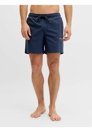 Mężczyzna w granatowych szortach kąpielowych ze sznurkiem, stojący boso na jednolitym jasnym tle.