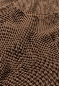 Hnědá pletená tkanina s ribbovanou strukturou, která zobrazuje vlnité vzory a různou těsnost v tkaní, zdůrazňující hloubku materiálu.