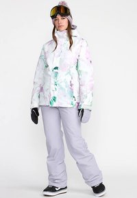 Veste de snowboard avec un imprimé floral pastel, fermeture éclair à l'avant et col montant. Associée à un pantalon de neige gris clair et des bottes de neige noires.