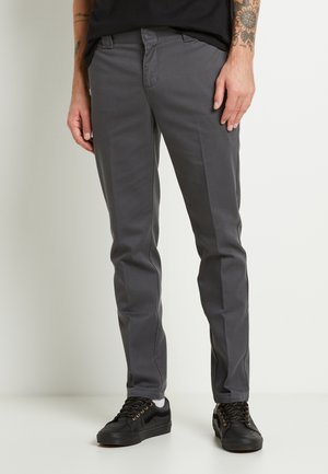 Mężczyzna ubrany w ciemnoszare, dopasowane spodnie oraz czarne trampki ze złotymi oczkami, stojący na tle jasnego, neutralnego tła.