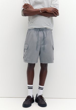 Osoba nosící šedé cargo šortky se stahovací šňůrkou, pruhované bílo-černé ponožky nad kotníky, tmavě hnědé šněrovací boty a bílé pruhované tričko s dlouhým rukávem, která má zkřížené ruce.