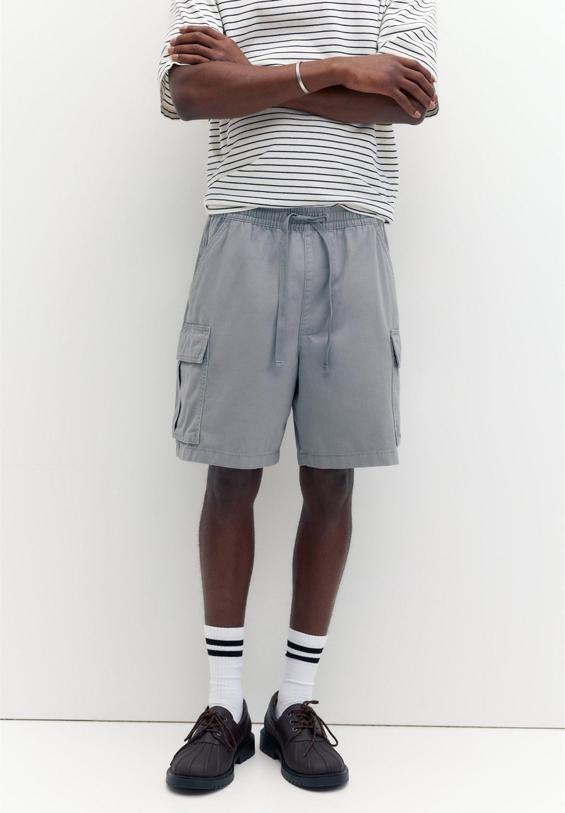 Personne portant un short cargo gris avec un cordon, des chaussettes blanches et noires rayées, des chaussures marron foncé à lacets et une chemise blanche rayée à manches longues, les bras croisés.