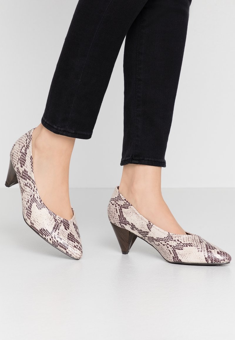 Deux pieds portant des chaussures à bout pointu, imprimé serpent, talons moyens, associées à un pantalon noir coupé, sur un sol gris clair.