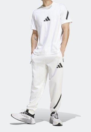 Mann trägt weiße Adidas-Sportkleidung mit Logo auf Shirt, Hose und schwarz-weißen Sportschuhen, steht mit den Händen in den Taschen.