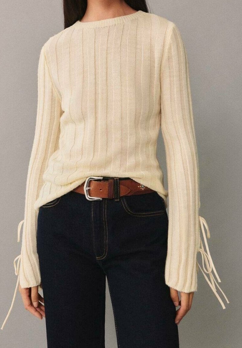 Femme portant un pull côtelé crème avec des cordons aux manches, rentré dans un jean foncé avec une ceinture marron et une boucle argentée.