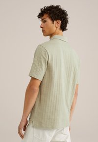 Camisa de manga corta de color verde pálido, hecha de una tela texturizada con un patrón sutil. Cuenta con un cuello clásico y aberturas laterales para mayor libertad de movimiento.