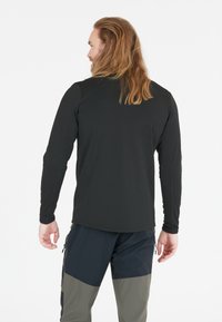 Sort langærmet top med tekstureret stof, tætsiddende pasform og flade sømme, båret med mørke flerfarvede bukser. Enkel design, ingen synlige logoer.