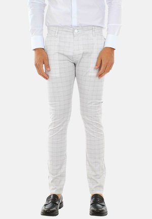 Uomo che indossa pantaloni attillati grigio chiaro a quadri, camicia bianca elegante e mocassini in pelle nera, in piedi contro uno sfondo bianco.