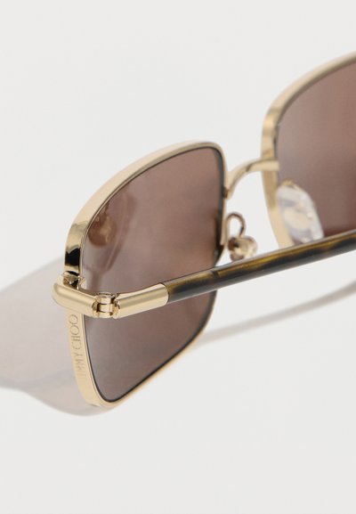 Γυαλιά ηλίου με χρυσή κορνίζα, καφετί φακούς με απόχρωση και λεπτό σκελετό, πάνω σε απλό λευκό φόντο.