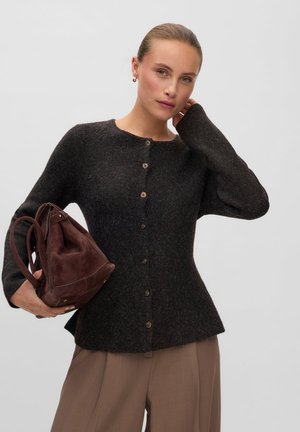 Femme portant un cardigan gris foncé boutonné et un pantalon beige, tenant un sac à main en cuir marron sur un fond clair uni.