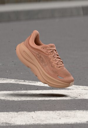 Ανοιχτό καφέ αθλητικό παπούτσι με παχιά σόλα, κορδόνια και διαπνέον δίχτυ, αιωρούμενο πάνω από άσφαλτο με λευκές ρίγες.