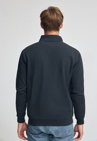 Felpa nera con design a colletto, maniche lunghe e polsi a costine, abbinata a jeans in denim blu chiaro. Mostrata vista posteriore.