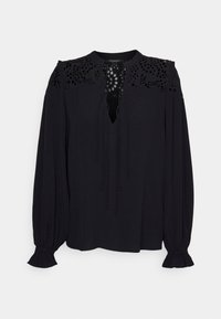 Blouse noire à manches longues avec détails en dentelle découpée sur le haut de la poitrine et les épaules, fermeture par lien au niveau de l’encolure et poignets volantés.