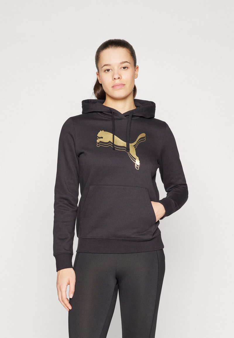 Crna dukserica s prednjim kengurujem, zlatni Puma logo. Ima kapuljaču sa vezicom i rebraste manžete. Uparena s crnim tajicama.