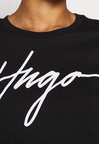 Lähiotteen kuva henkilöstä, joka pitää päällään mustaa paitaa, jossa on valkoinen käsin kirjoitettu logo, joka alkaa sanalla "Hugo" rinnan kohdalla.