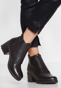 Botas de cuero negras hasta el tobillo con punta redonda, cremallera lateral, acentos texturizados y un tacón apilado, colocadas sobre pantalones negros con cremalleras.