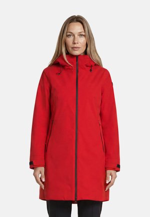 Femme portant un long manteau rouge à capuche avec fermeture éclair frontale et poches latérales zippées, debout devant un fond uni.