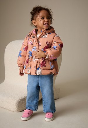 Lächelndes Kleinkind trägt eine bunte Steppjacke mit Tierdruck, blaue Jeans und pinke Sneakers, steht neben einem beigen, strukturierten Stuhl.